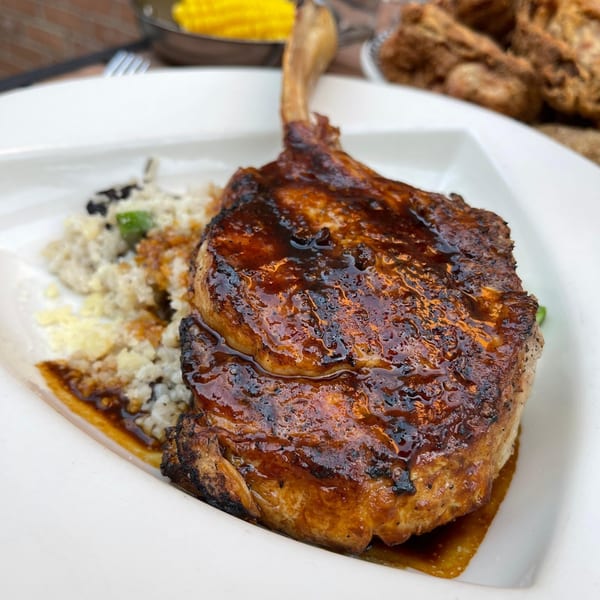 A glistening, bone-in pork chop with a dark glaze, served with a side of rice and sauce.