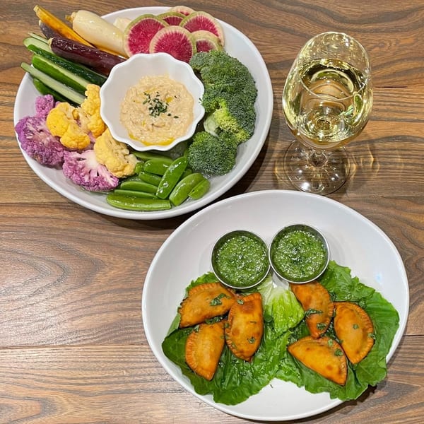 Plates of colorful crudités with dip, empanadas with green sauce, and a glass of white wine.